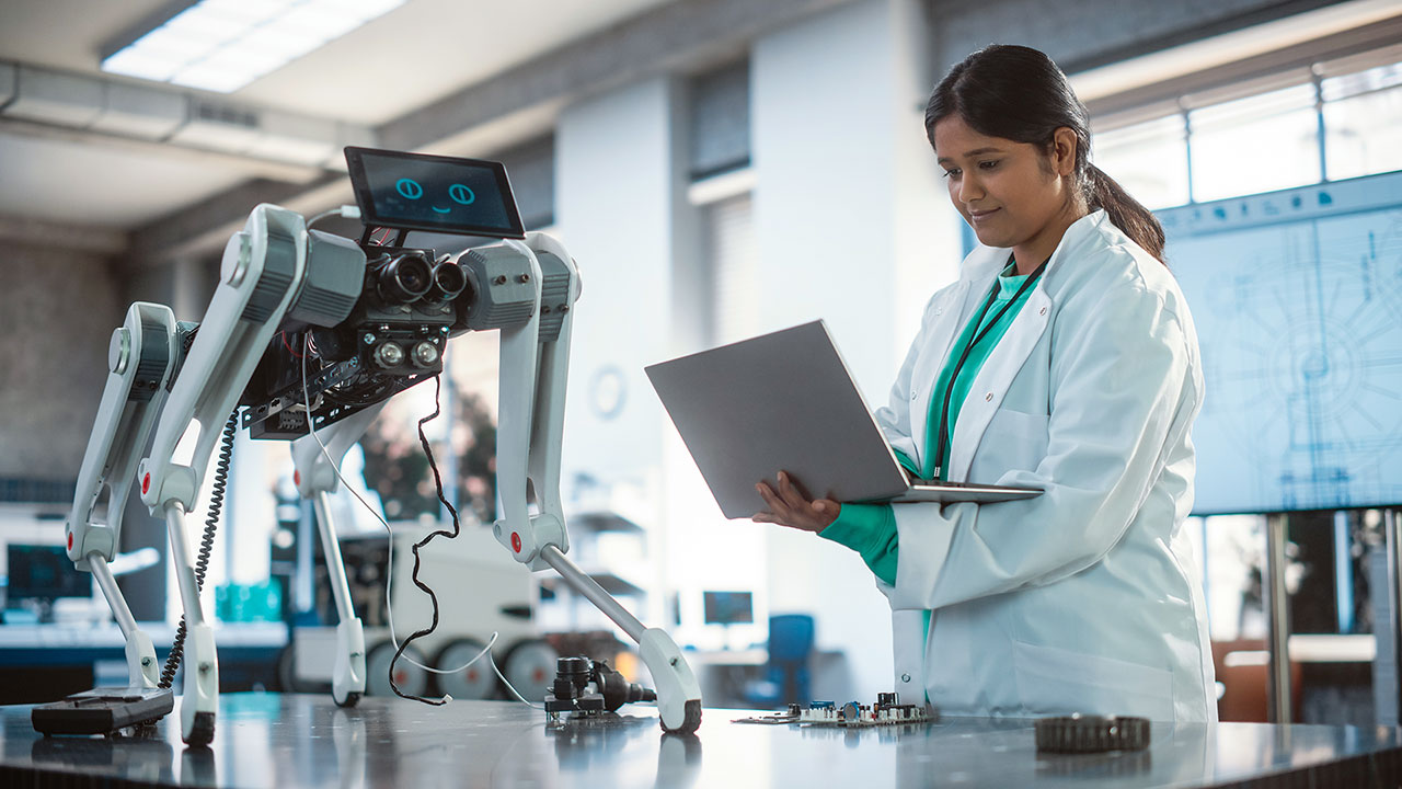 A woman in a lab coat works on a laptop while standing next to a futuristic, four-legged robotic machine with a screen displaying animated eyes. The robot, equipped with cameras, sensors, and exposed wiring, stands on a metallic table alongside scattered electronic components and tools. The background shows a bright, modern lab with diagrams displayed on a screen, creating an atmosphere of advanced technology and innovation. The woman appears focused and engaged, suggesting a collaborative effort in robotics or AI development.