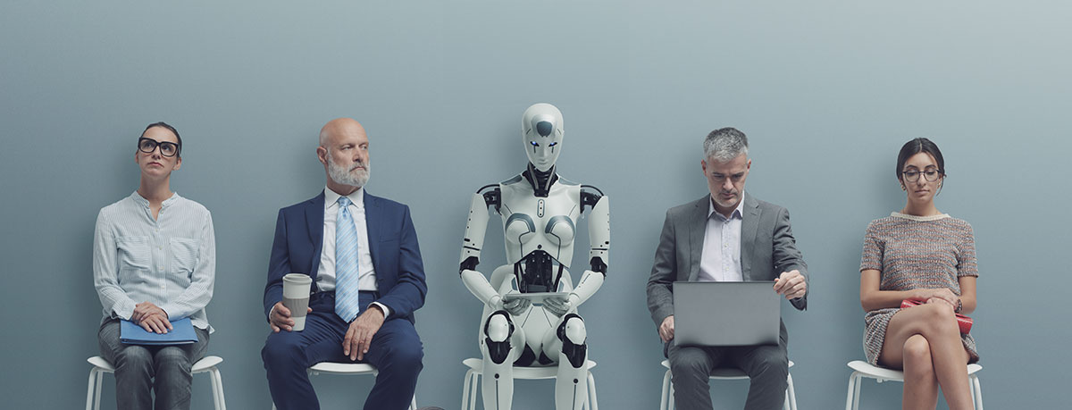 Four human beings sit in a row of white chairs up against a wall waiting. In the center chair sits a futuristic-looking full-bodied robot typing on a laptop. The robot also appears to be waiting along with everyone else.